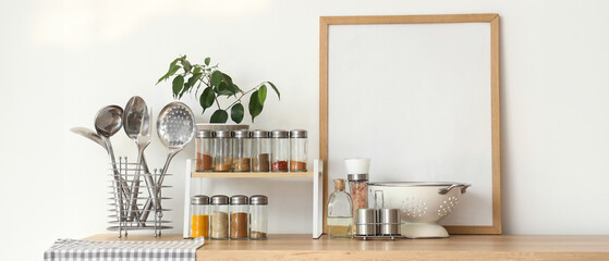 Shelf with different spices, plant, blank frame and utensils on counter near light wall in kitchen