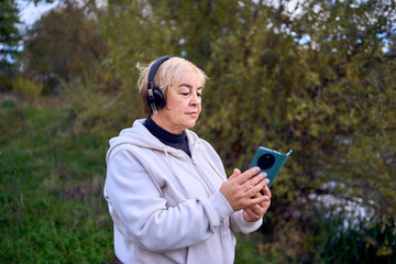 elderly woman switches the music in her wireless headphones using her phone during a morning run in...