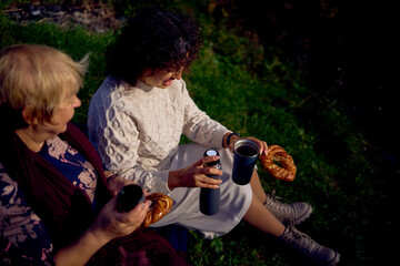 middle-aged daughter and old mother eating pastries and drinking tea from thermos cups on a picnic in the autumn forest