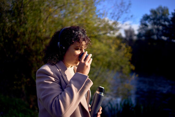 middle-aged woman in wireless headphones drinking coffee, tea from a thermos on a walk in the autumn forest