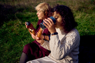 middle-aged daughter and old mother eating pastries and drinking tea from thermos cups on a picnic in the autumn forest