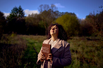 a middle-aged woman in a beige coat with a holy bible in her hands prays for peace in the autumn...