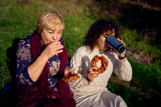middle-aged daughter and old mother eating pastries and drinking tea from thermos cups on a picnic in the autumn forest