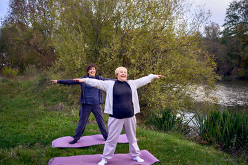 middle-aged woman teaches her old mother somatic exercises and yoga to maintain health by the river in autumn
