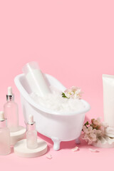 Composition with mini bathtub, bottles of cosmetic products and flowers on pink background