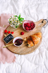 Süsses Frühstück mit Croissant und Himbeerkonfitüre auf einem Serviertablet auf einer Bettdecke. 