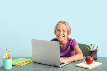 Little boy with laptop doing homework at table near blue wall