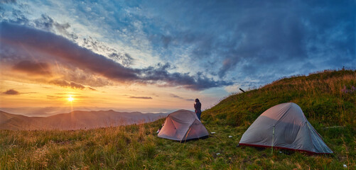 A camper stands beside tents pitched on a grassy mountain slope, gazing at a glowing sunset over distant ridges under a dramatic sky filled with streaked clouds and fading golden light.
