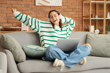 Beautiful young happy woman in headphones with laptop resting on sofa at home