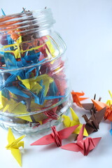 A vibrant mix of colorful origami cranes inside a clear glass jar with several scattered around it,...