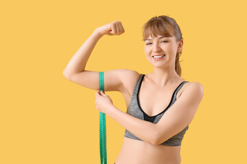 Beautiful young sporty woman measuring her biceps on yellow background. Weight loss concept
