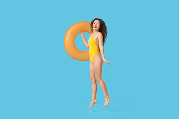 Jumping young Asian woman in stylish yellow swimsuit with inflatable ring on blue background