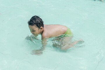 A kid with curly hair is in a swimming pool, wearing green swim trunks. The child is on all fours,...
