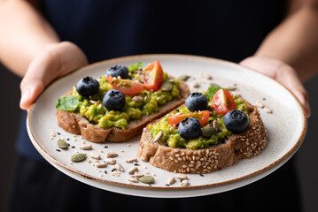 Avocado toast with blueberries, cherry tomatoes, sunflower seeds, pumpkin seeds, sesame seeds and other grain on plate holding by hand, Healthy eating