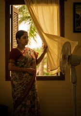 An Indian woman standing by a window with a fan, lifting the curtain to catch air. Trying to find relief from hot summer weather. Concept of home cooling and comfort.