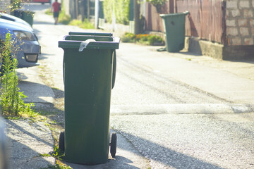 Ecological habits and suburban life: green trash bins standing on a residential suburban street on a sunny summer day. Concept of sanitation service, eco habits and community waste sorting culture
