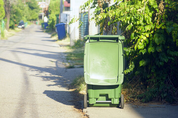 Waste collection and sorting in the everyday life of a small European city: a green wheeled trash bin with an open lid on a deserted sunny street and blue garbage containers in the distance.