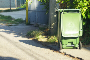 Green plastic trash bin standing on asphalt street near fence. Symbolizing urban waste collection, eco-conscious recycling habits and sanitation infrastructure in quiet small-town residential areas