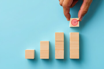 Achieving Goals A Hand Places the Final Wooden Block with a Target Symbol on Top of a Rising Bar Graph Against a Light Blue Background in a Studio Shot