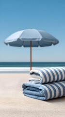 Striped beach towels neatly stacked on sandy shore, with a blue umbrella providing shade, creating a relaxing summer atmosphere by the ocean waves and clear sky