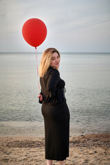 Young woman in elegant black attire stands on tranquil beach, holding red balloon. Peaceful...