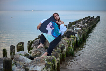 Young female artist peacefully holds her painting while sitting on coastal rocks. Tranquil seascape...