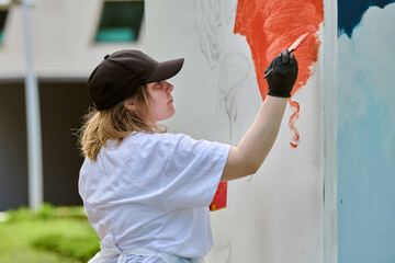 Young adult female artist, with light hair and casual attire, paints vivid mural. Outdoors setting with green space. Bright colors and focused expression © TRAVELARIUM