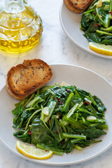 Cooked chicory greens sautéed in olive oil, garlic, and hot pepper flakes - a traditional Italian side dish served on a plate with toasted bread and a slice lemon. A bottle of olive oil on the side.