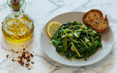 Sauteed Chicory Greens - a traditional Italian side dish on a plate served with toasted bread and a slice of lemon. A bottle of olive oil and some scattered hot pepper flakes on the side.