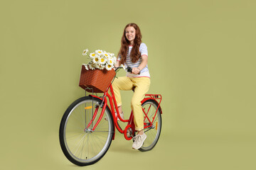 Smiling teenage girl riding bicycle with basket of flowers against green background