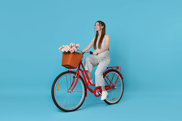 Smiling woman riding bicycle with basket of flowers against light blue background
