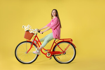 Smiling woman riding bicycle with basket of flowers against light green background