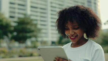 Portrait African American lady looking at tablet computer screen against multistorey building. Female surfs internet via device standing on summer city street - Powered by Adobe