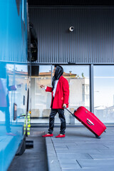 Stylish non-binary person leaving bus station after trip with red suitcase