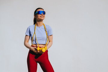 A joyful woman, sporting futuristic sunglasses, confidently holds yellow dumbbells against a clean,...