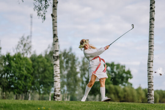 Young woman in traditional attire and flower crown swinging a golf club on a green field surrounded by birch trees during daylight.