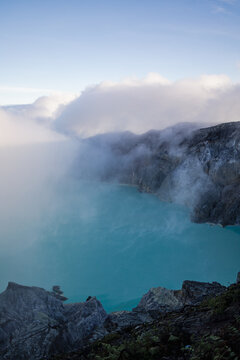 View of a turquoise lake nestled within a volcanic crater, shrouded in ethereal mist and framed by rugged, dark rocks, Ijen Volcano Crater, East Java, Indonesia.
