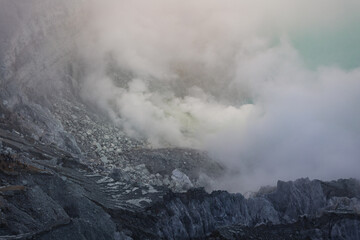 View of ethereal clouds swirling around the rugged, textured terrain, revealing a hint of turquoise waters, creating a mystical landscape, Ijen Volcano Crater, East Java, Indonesia.