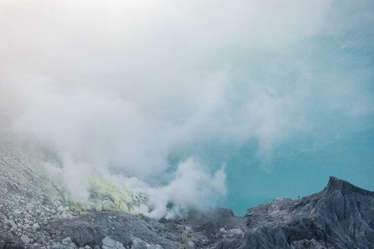 View of sulfuric smoke billowing from the volcanic vents over jagged, rocky terrain against a pale blue sky, Ijen Volcano Crater, East Java, Indonesia.