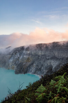 View of a breathtaking scene where turquoise waters meet rugged cliffs shrouded in mist, under a pastel sky, Ijen Volcano Crater, East Java, Indonesia.