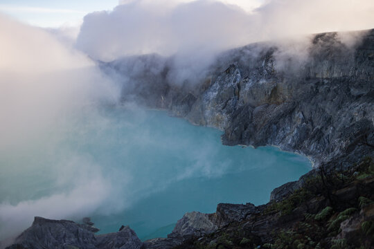 View of an otherworldly scene, where a turquoise lake meets jagged rocks amidst swirling clouds, creating an ethereal landscape, Ijen Volcano Crater, East Java, Indonesia.