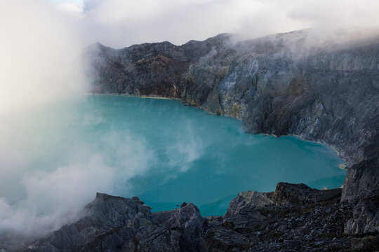 View of a vibrant turquoise lake nestled within the rugged crater, shrouded in ethereal clouds and dramatic rocky terrain, Ijen Volcano Crater, East Java, Indonesia.