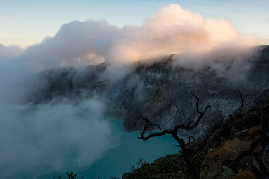 View of a turquoise crater lake nestled beneath swirling clouds and rugged volcanic cliffs, framed by stark, bare branches, Ijen Volcano Crater, East Java, Indonesia.