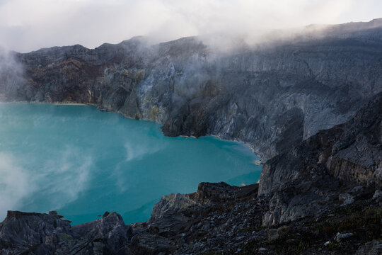 View of the serene yet acidic turquoise lake nestled within the rugged, volcanic crater shrouded in mist, a stark contrast of colors and textures, Ijen Volcano Crater, East Java, Indonesia.