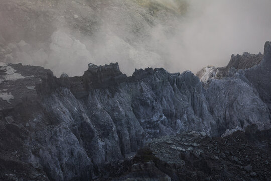 View of rugged, grey volcanic rock formations shrouded in swirling white smoke, creating an otherworldly and dramatic landscape, Ijen Volcano Crater, East Java, Indonesia.
