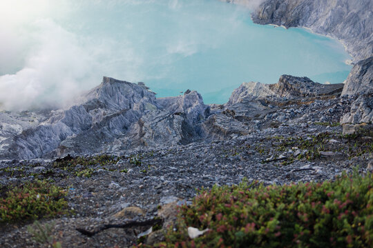 View of the rugged, rocky landscape contrasts with the ethereal turquoise lake and rising steam, creating a surreal scene, Ijen Volcano Crater, East Java, Indonesia.