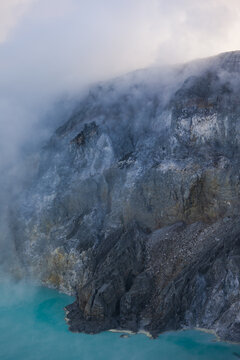 View of the rugged, smoky terrain contrasts with the turquoise lake below, shrouded in a ghostly mist, Ijen Volcano Crater, East Java, Indonesia.