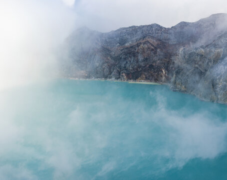 View of the turquoise lake contrasting with the rugged dark volcanic rock and swirling mists creating an ethereal panorama, Ijen Volcano Crater, East Java, Indonesia.