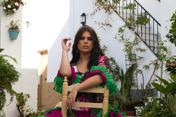 Young and beautiful woman dancing flamenco sitting on an old wooden chair in a typical street of andalusia. She is dressed in a tailed gown with ruffles and flowers. The woman is a flamenco dancer.