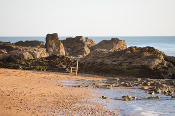 An old wooden chair on the shore of the beach next to the rocks.